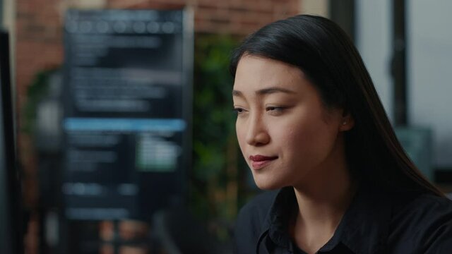 Focused asian software developer writing programming code and smiling sitting at desk in database development agency. Friendly coder computing big data casually in it startup office.