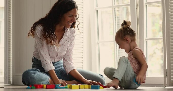 Cheery mom play wooden multicoloured bricks with preschool 4s daughter, sit on warm floor inside cozy room build tower laughing enjoy carefree leisure at home. Fun, developmental games of kids concept