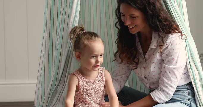 Cute girl play with loving Latina mommy or babysitter sit under cloth canopy in playroom. Happy family enjoy leisure, have fun, communicate spend time together. Playtime at home with children concept