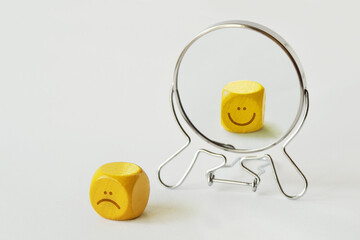 Yellow dice with sad expression looking in the mirror and seeing himself smiling - Concept of person who hides his pain behind his smile