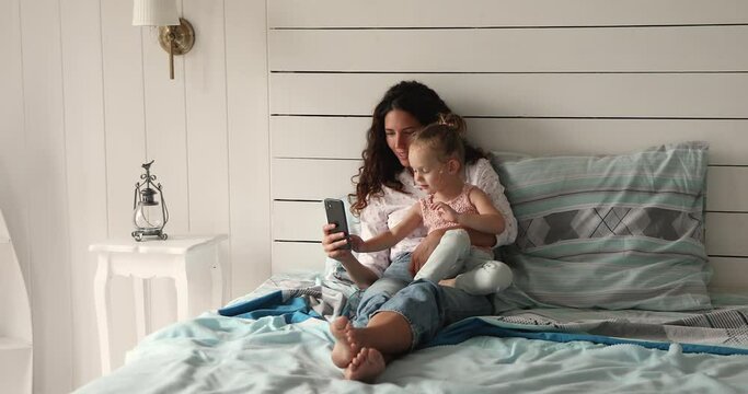 Latina woman and little 4s girl rest on bed in bedroom using smartphone having fun enjoy new mobile application, play educational developmental on-line games, spend leisure use wireless tech concept