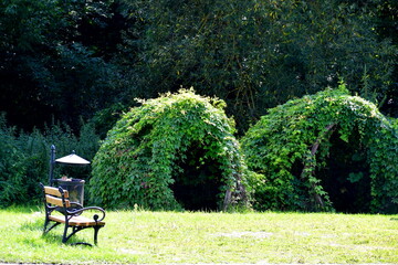 A view of a section of a public park with some wooden bench allowing for rest and shelter or huts for animals made out of vines, planks, boards, and branches of trees seen in pPoland on a summer day