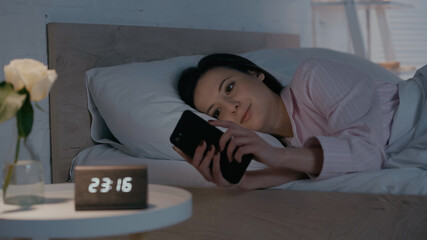 Brunette woman using smartphone on bed near blurred clock in bedroom.