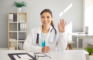 Webcam view head shot portrait of happy smiling Caucasian female doctor in white lab coat sitting at table and waving hello greeting patient at online consultation via video call. Telemedicine concept © Studio Romantic