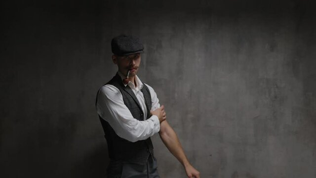 Young dangerous gangster man dressed in vintage suit, cap and with smoking pipe in his mouth enters, rolls up his sleeves, prepares for a fight and stands in fighting stance in front of concrete wall