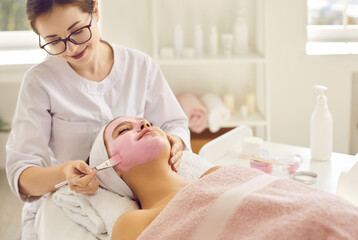 Obraz na płótnie Canvas Happy woman getting facial treatment done by beautician in beauty salon or spa center. Beautiful young lady relaxing under soft towel and enjoying pink clay face mask for clear fresh rejuvenated skin