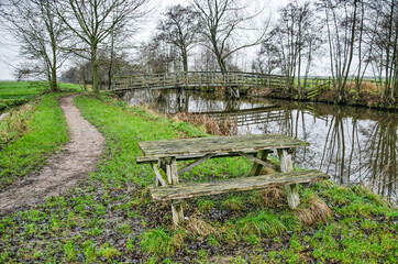Fototapeta na wymiar Wooden bench, bridge across a canal and muddy hiking trail in Krimpenerwaard polder in the Netherlands