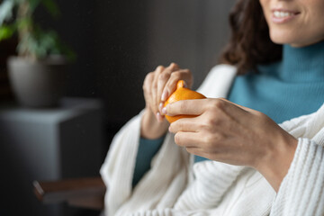 Obraz na płótnie Canvas Smiling woman peels juicy ripe sicilian tangerine at home, selective focus on hands. Happy female wrapped in white knitted plaid eating seasonal winter fruits, defocused. High vitamin C food concept