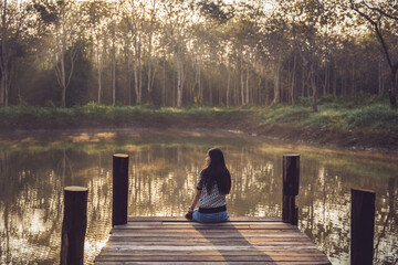 A sad woman sits floating on an old wooden bridge that juts out into a pond in nature. Lonely sad...