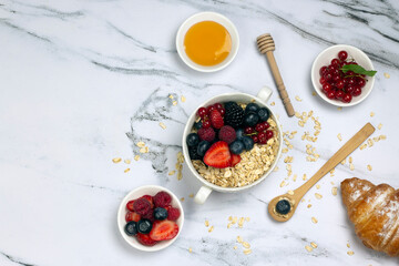 Healthy breakfast, oat with berry, honey, croissant and ripe sweet berries on white marble table. top wave, flat lay.