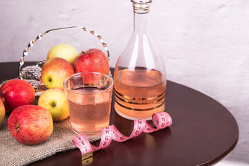 On a wooden table there is a glass and a decanter with apple cider vinegar and a plate with apples.
