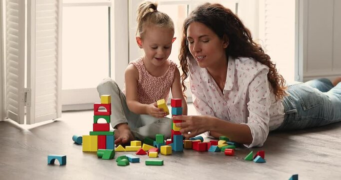 Young Latina mother play multi colored wooden cubes with cute little 4s daughter, lying sit on warm floor with underfloor heat system enjoy playtime at comfort home. Fun, communication, growth concept