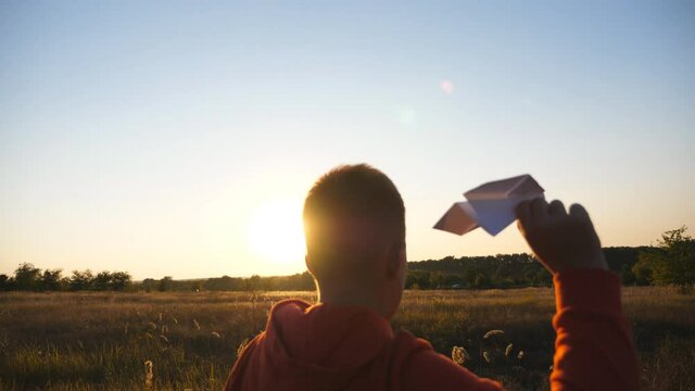 Carefree little child jogging with a paper airplane through grass field. Small boy running with toy plane among meadow and launching toy plane over sunset background. Concept of childhood dream