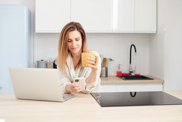 young woman using laptop and drinking a coffee while sitting at home kitchen.