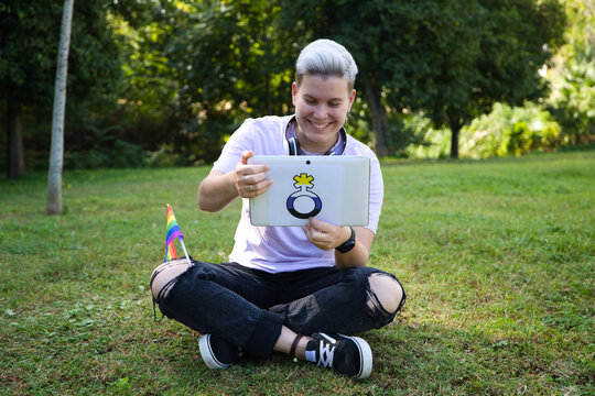 non-binary person sitting in the park with a tablet with the non-binary symbol and the gay pride flag on his knee. Diversity and gay pride. Non-binary visibility concept.