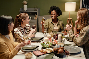 Group of young friends sitting at festive table drinking alcohol drinks and celebrating holiday together at home