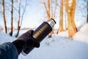 The hand holds a thermos against the background of nature. Black metal thermos for tea. Keep warm in winter. Tourist utensils.