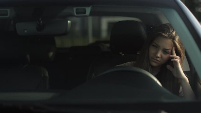 A tired woman is sitting in her car in the driver's seat. She is leaning against the door and waving her head. The sun is shining on her. 4K