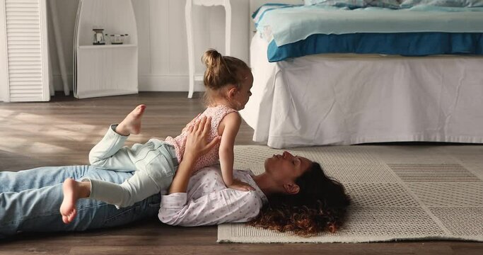 Young affectionate Latina mother touch noses play with little cute 4s daughter lying on warm floor in cozy bedroom at home, having fun enjoy moments of tenderness and sweet time together feeling love