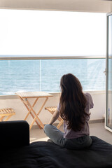 A young woman is sitting on sofa near balcony and looking at the sea