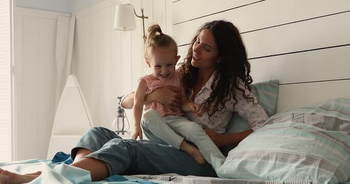 Cheery Hispanic mother play with cute little 4s daughter, sit on bed in warm bedroom laughing, enjoy playtime together at home. Happy motherhood, babysitting, custody and adoption, childcare concept
