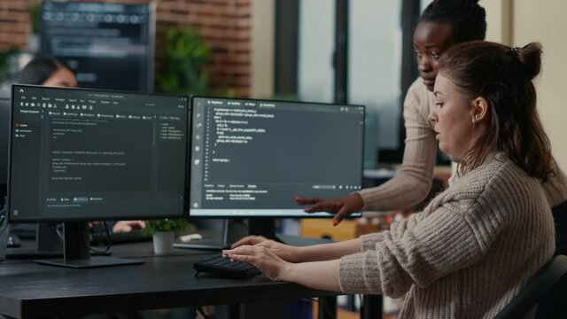 Junior programer writing algorithm under experienced colleague supervision sitting at desk with multiple screens showing programming interface. Developer writing code with the help of colleague.