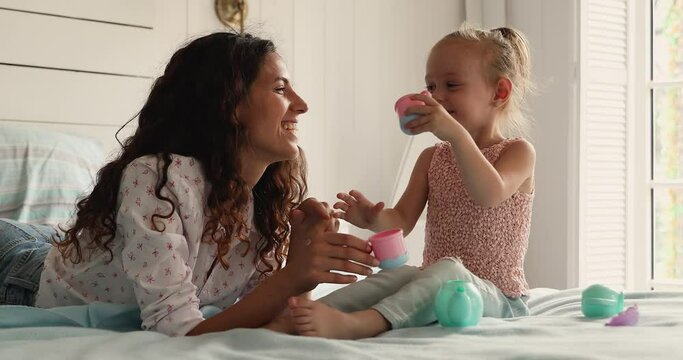 Hispanic mom her 4s daughter holds teacups play tea party have fun fooling relax on bed in cozy bedroom, laughing enjoy playtime use playthings spend leisure at home. Childhood girlish games concept