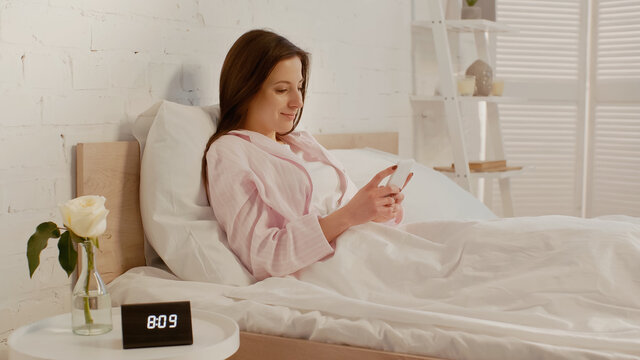 Happy woman using smartphone on bed near clock and flower on bedside table.