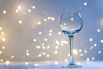 Transparent wine glass, with a bokeh of bright lights. A wine glass on a light background, in a cold key, with a garland side. A postcard for holidays and dinners.