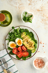 Fototapeta na wymiar Avocado, cherry tomato, spinach and chicken egg, microgreens peas and black sesame seeds fresh salad in bowl on white stone table background. Healthy breakfast food concept. Top view.