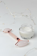 Concept of winter skin care. Closeup of a jar of moisturizer and a rose quartz roller with fairy lights in the background.