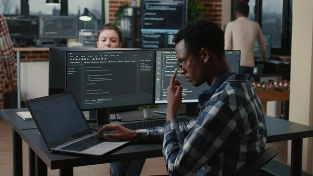 Focused programer fixing glasses while typing on laptop sitting at desk with multiple screens parsing code. Coder working on user interface with team of programmers coding big data in the background.