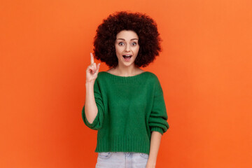 Happy surprised woman with Afro hairstyle wearing green casual sweater raising finger up, has sudden great idea, looking at camera with open mouth. Indoor studio shot isolated on orange background.