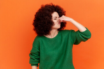 Portrait of young adult woman with Afro hairstyle wearing green casual style sweater holding breath with fingers on nose, stinky odor, fart. Indoor studio shot isolated on orange background.