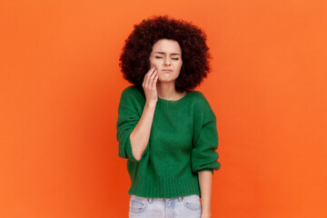 Sick woman with Afro hairstyle wearing green casual style sweater touching her cheek, suffering...