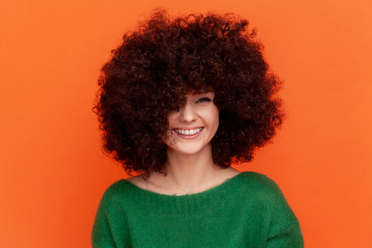 Smiling woman with Afro hairstyle wearing green casual style sweater looking at camera, showing her fluffy hair, advertising beauty salon. Indoor studio shot isolated on orange background.