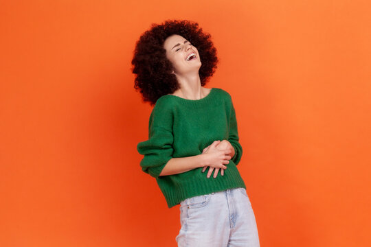 Happy woman with Afro hairstyle wearing green casual style sweater holding belly, laughing, hearing funny joke or anecdote, being in good mood. Indoor studio shot isolated on orange background.
