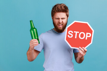 Portrait of bearded man showing alcoholic beverage beer bottle and stop sign, warning and worrying,...