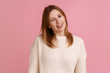 Fototapeta premium Portrait of positive optimistic blond woman standing and winking playfully, positive expression, looking at camera, wearing white sweater. Indoor studio shot isolated on pink background.