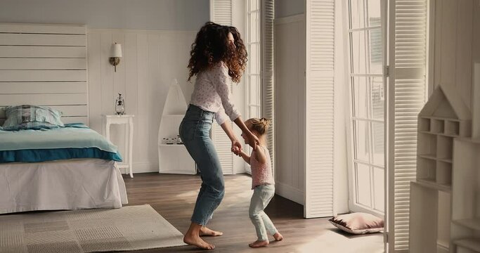 Latina woman her cute adorable little 4s daughter jump barefoot on warm floor listen favourite song have fun holds hands dancing in cozy bedroom at modern home. Family pastime, leisure, hobby concept