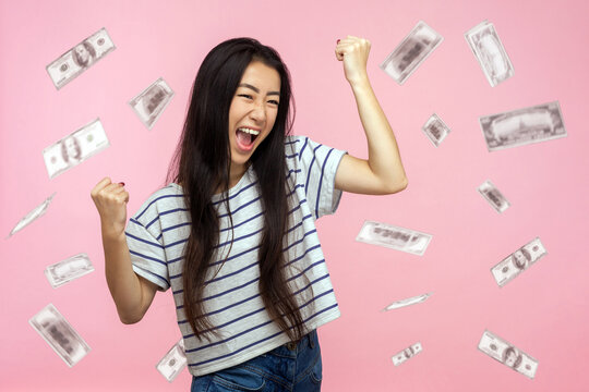 Money rain, winner and rich. happy young ecstatic motivated woman standing with raised fists and shouting for joy, winner excited for success. dollars falling. indoor isolated on pink background