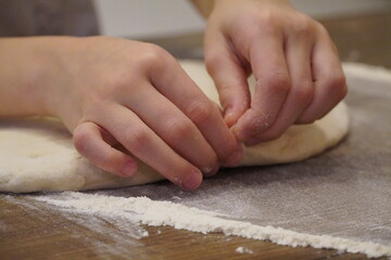 Young hands pinch and crumple the dough on a brown wooden table sprinkled with flour.