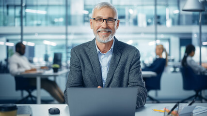 Modern Office: Portrait of Successful Middle Aged Bearded Corporate Businessman Working on a Laptop at his Desk, Looking at Camera, Smiling. Diverse Workplace with Professionals. Front View Shot