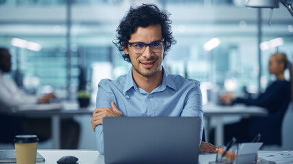 Modern Office Businessman Working on Computer, Smiling, Looking at Camera. Portrait of Successful Latin IT Software Engineer Working on a Laptop at his Desk. Front View Shot