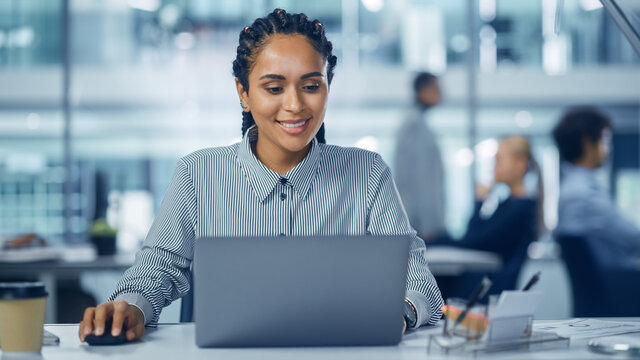 Multi-Ethnic Office: Professional Black Female IT Engineer Working on Desktop on Laptop Computer. Successful, Smiling Businesswoman with Braided Hair Does Creative Job. Confident Product Designer