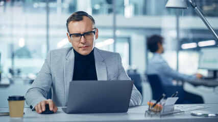 Modern Office: Portrait of Successful Middle Aged Businessman Wearing Suit and Glasses Working on a Laptop at his Desk. Serious, Confident, Stylish Corporate Executive Using Computer. Front View Shot