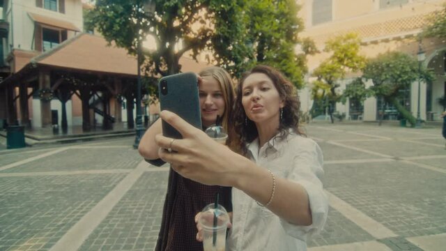 Two young smiling girls take selfie photos on a smartphone. Models pose in front of the street.