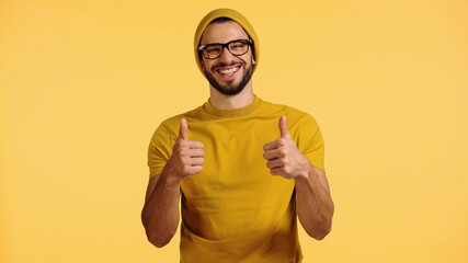 happy young man in beanie hat, glasses and t-shirt showing thumbs up isolated on yellow