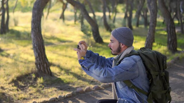 Young man taking landscape photograph in the forest with his smart phone. He wears a hat, a denim jacket and a mountain backpack.