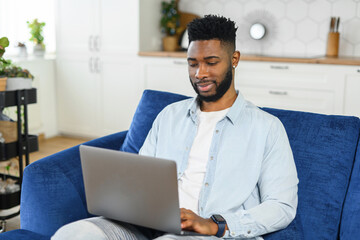 Smiling African-American freelancer guy is using laptop computer sitting on the couch at home, hindu guy enjoys remote work from home in comfy atmosphere. Black man spends leisure online